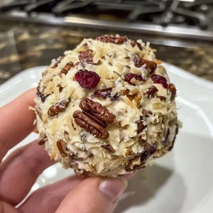 ['Side view of a cranberry pecan cheese ball garnished with nuts and chives.', 'Close-up of a delicious cheese ball made with cranberries and pecans on a wooden serving board.', 'A colorful cranberry pecan cheese ball, featuring chopped nuts and herbs, ready to be served.', 'Juicy cheese ball with a textured surface of dried cranberries and toasted pecans.']